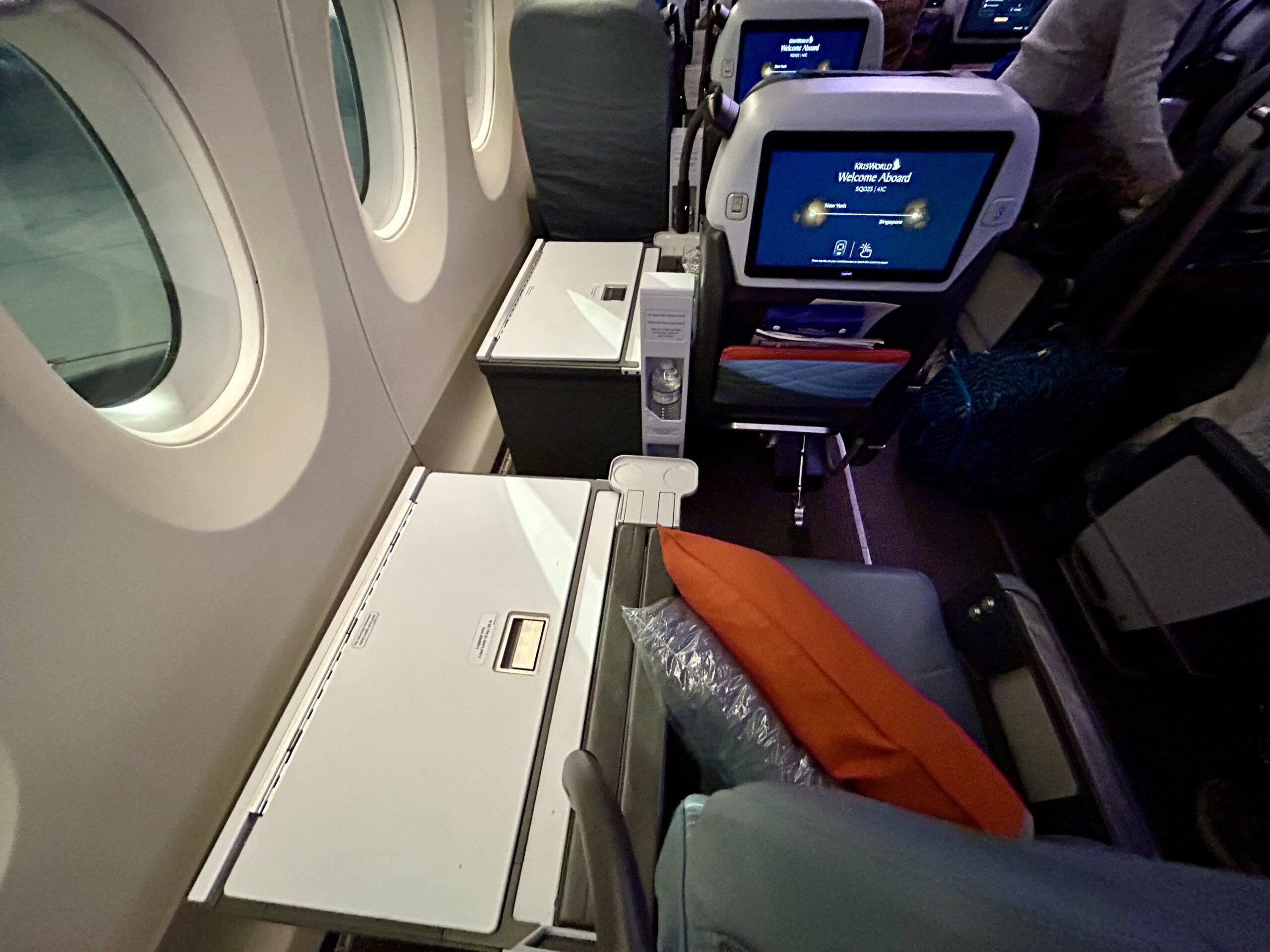 airplane seat with an orange pillow, armrest and tv screen